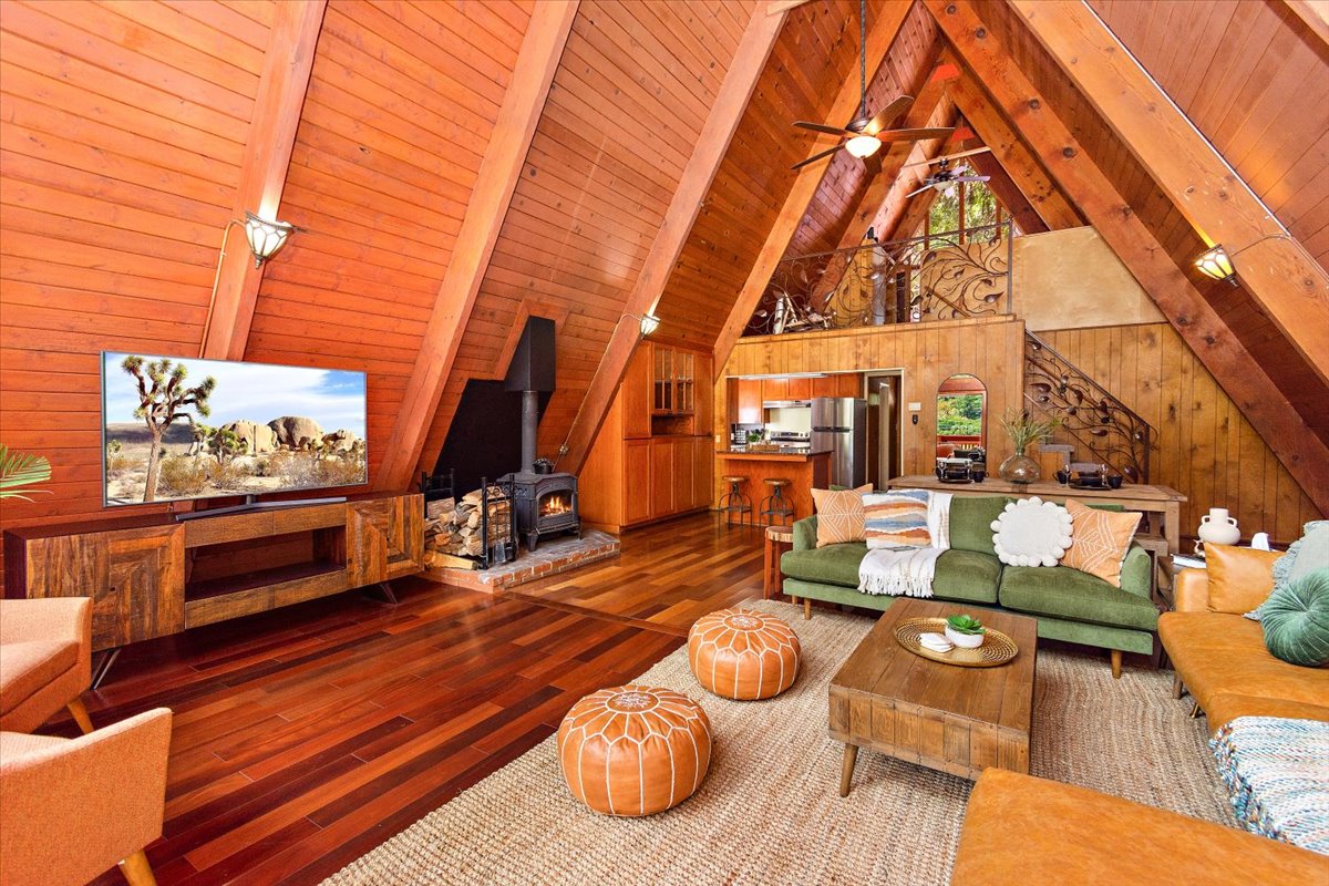 A spacious living area features high wooden ceilings with exposed beams. A large flat-screen television is mounted on the wall, while a cozy fireplace sits nearby. Comfortable seating includes a green sofa, a wooden coffee table, and poufs, enhancing the inviting atmosphere of the room.