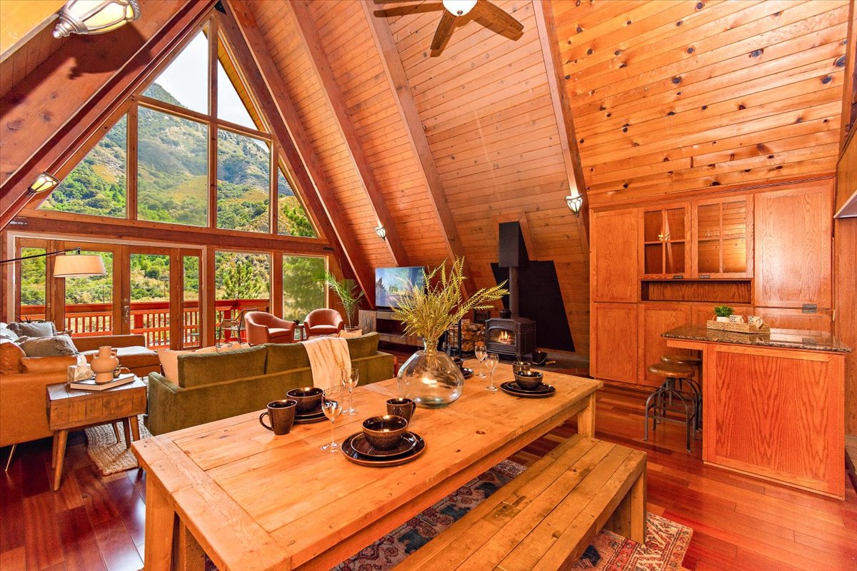 An inviting open living space is highlighted by vaulted wooden ceilings and large windows that frame views of the surrounding mountains. A wooden dining table is set for a meal, complemented by a cozy seating area with a fireplace and natural light filtering through the expansive glass.