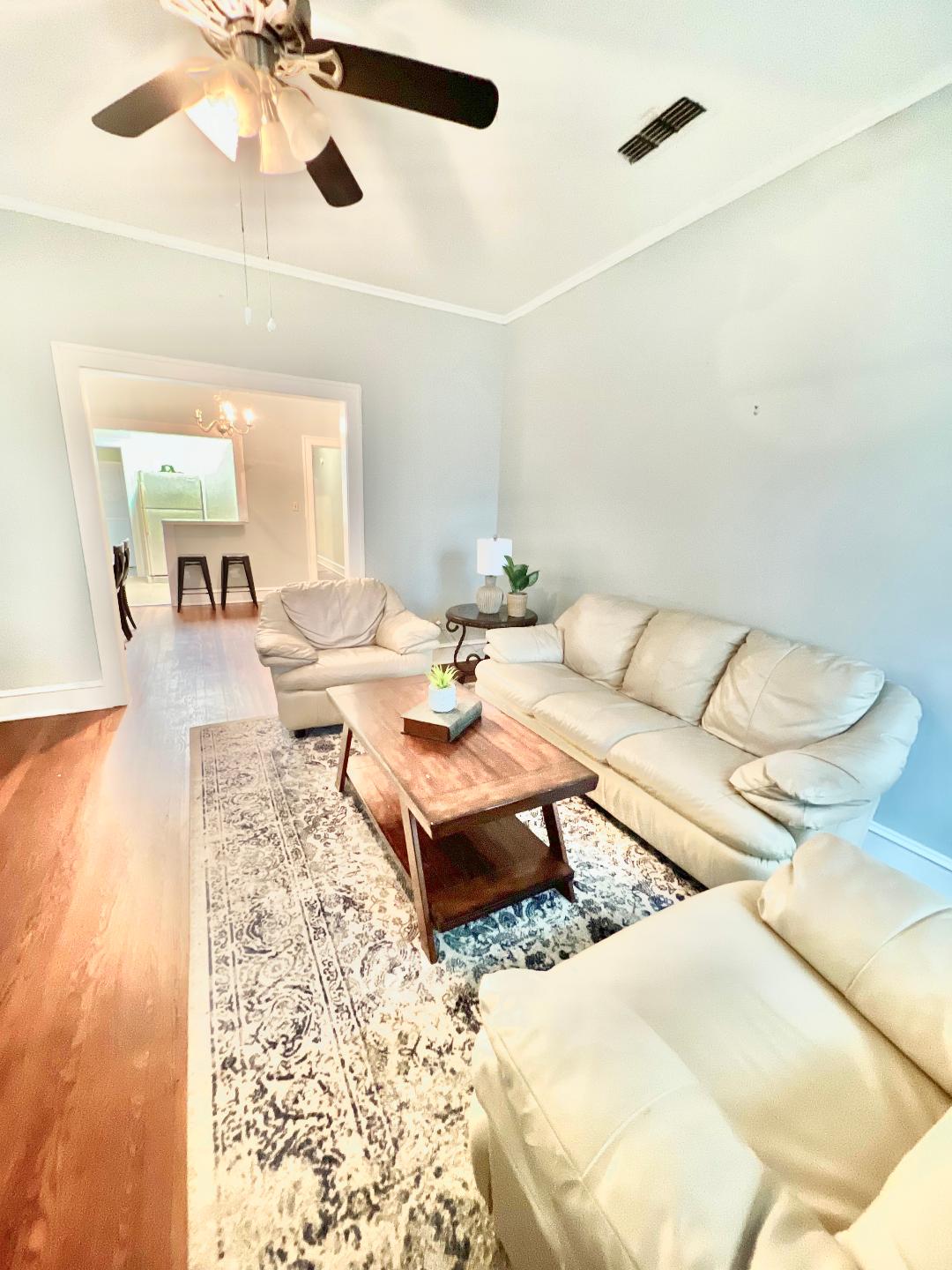A living area features light-colored leather sofas surrounding a wooden coffee table. A decorative plant sits atop the table, and a patterned area rug adds texture. A ceiling fan overhead enhances airflow, while natural light enters from an adjacent room.