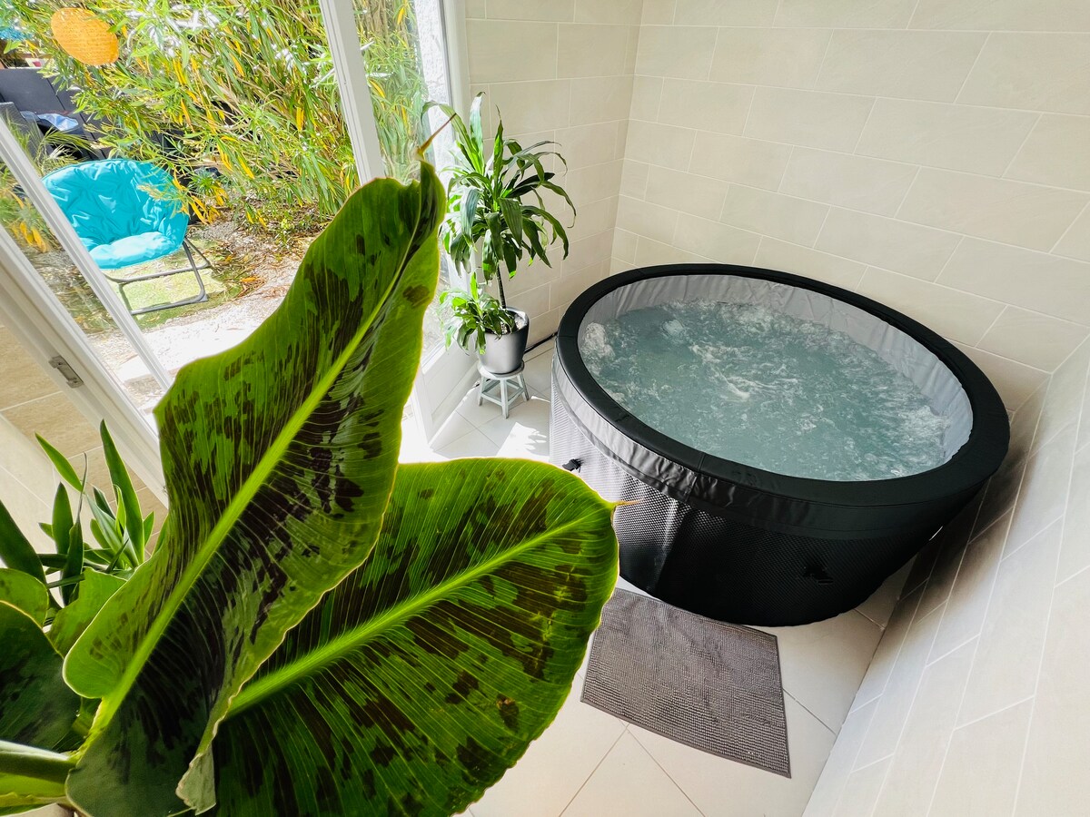 A private spa area contains a round, inflatable hot tub filled with bubbling water. The space is adorned with potted plants, adding a touch of greenery. Natural light floods the room through large glass doors, creating a serene atmosphere.