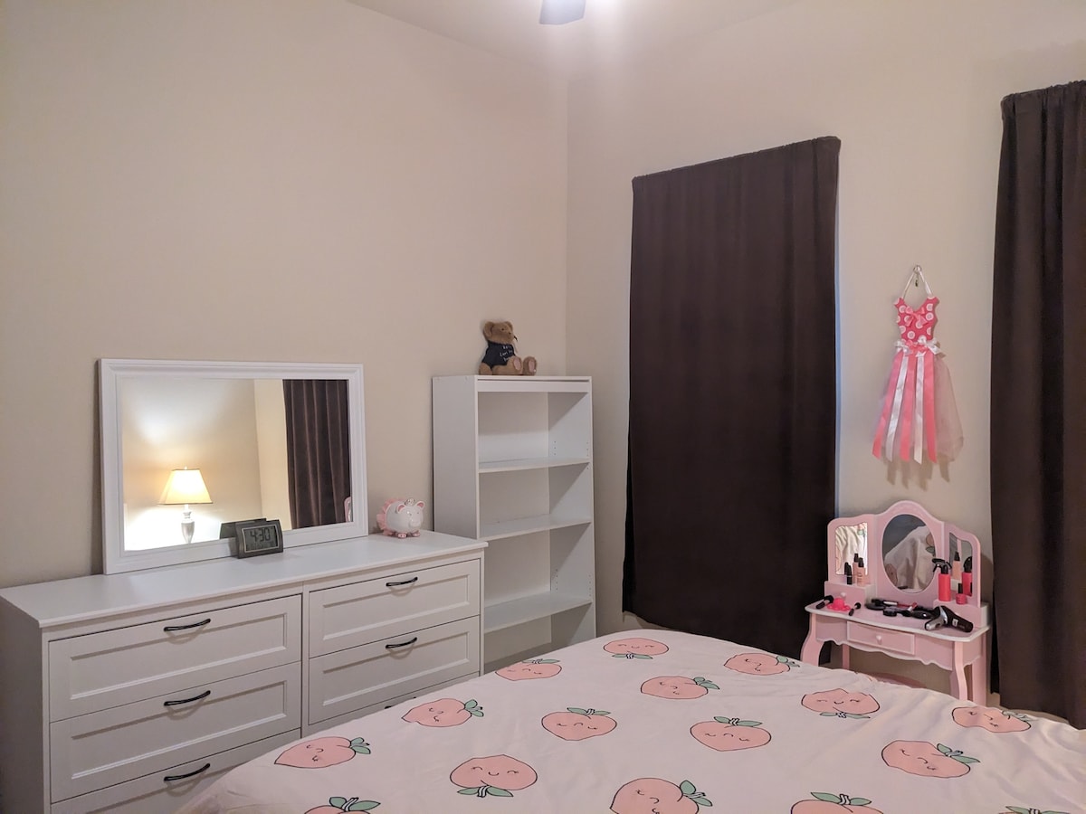 A cozy bedroom features a queen-sized bed adorned with a patterned duvet cover. A white dresser is positioned against one wall, complemented by a playful vanity set adorned with cosmetics. Two dark curtains frame a window, and a bedside lamp provides soft illumination.