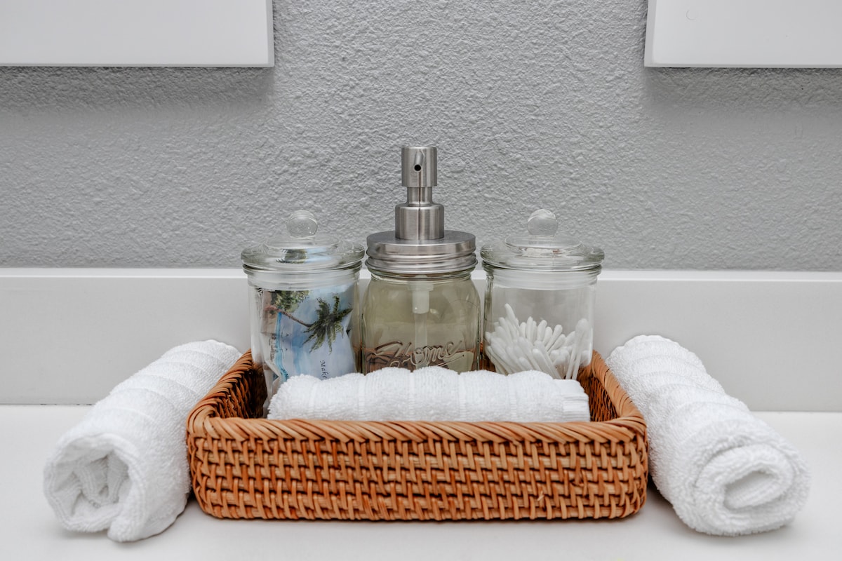 A woven basket holds neatly arranged toiletries, including glass jars with cotton swabs and a scented spray. Fresh white towels are rolled and placed beside the basket, creating a tidy and organized bathroom setup.