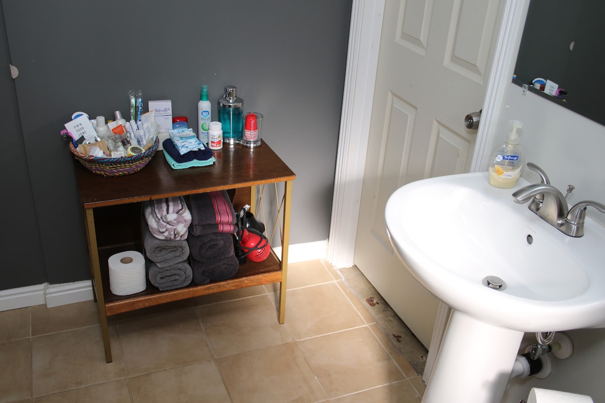A neatly organized bathroom is presented, featuring a wooden shelf stocked with toiletries and a variety of personal care items. A porcelain sink is visible alongside a bathroom door. Soft towels are neatly stacked, with a roll of toilet paper placed below.