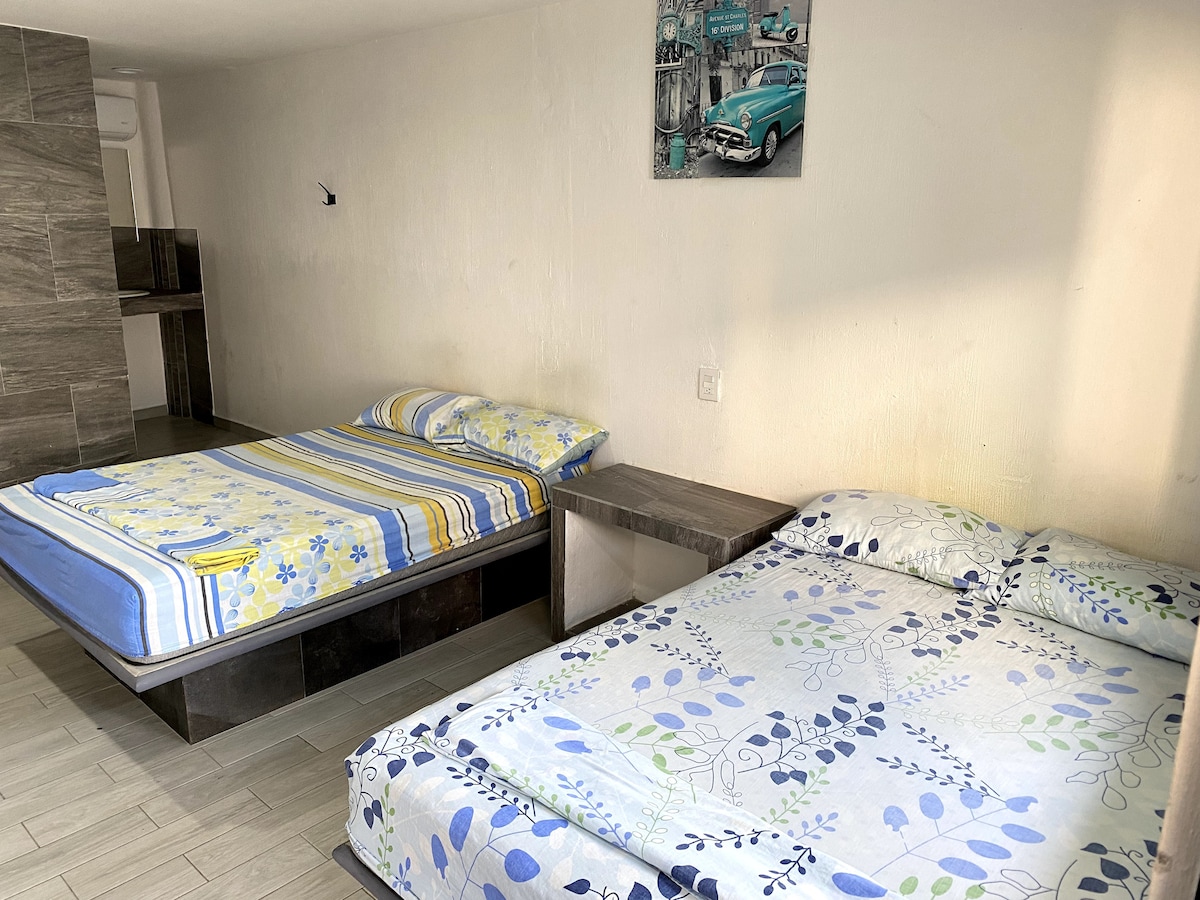 A cozy room is equipped with two spacious beds dressed in floral and striped bedding. A small side table sits between the beds. Light from a nearby window softly illuminates the room, highlighting the neutral-toned walls and floor.
