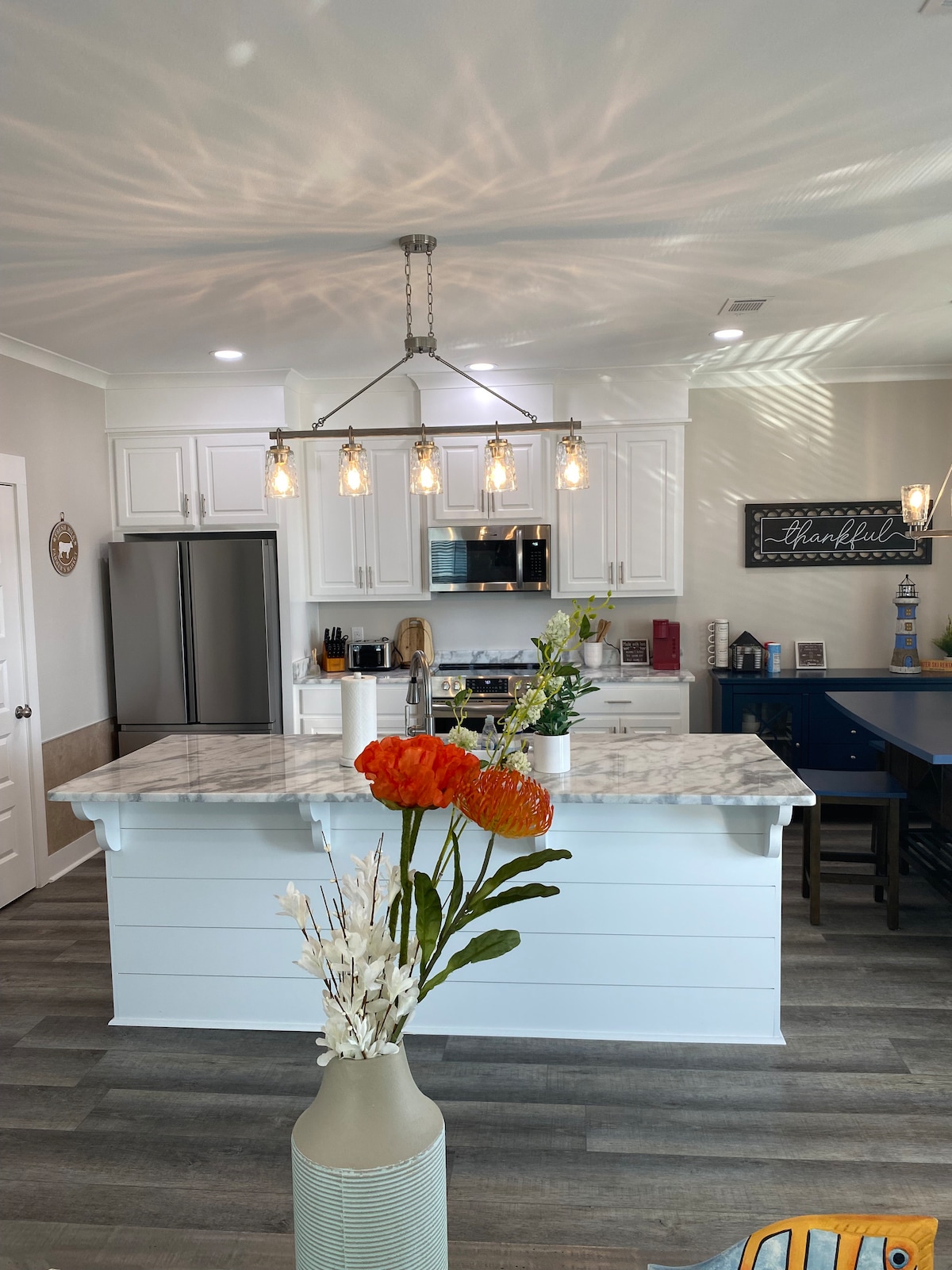A modern kitchen features white cabinetry and a marbled countertop on a spacious island. Pendant lights hang above, illuminating the area. A vase with bright flowers adds a pop of color, while stainless steel appliances complete the contemporary design.