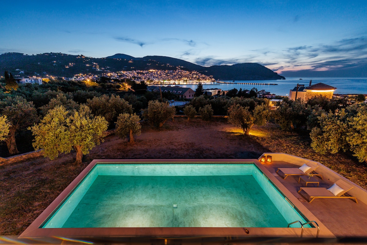 The image displays a tranquil swimming pool illuminated at night, with a surrounding area of olive trees. In the backdrop, the charming town of Skopelos and its harbor are visible, glimmering under the evening sky. Soft lighting enhances the serene atmosphere.