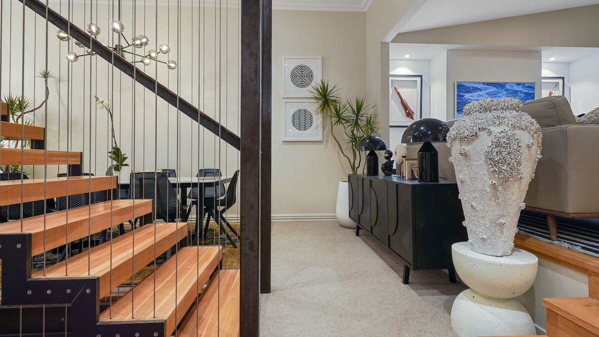 A stylish entryway is highlighted by a wooden staircase with a modern railing. A dining area is visible in the background, complemented by a decorative plant. Textured decor, including a sculptural vessel and sleek furnishings, adds character to the space.
