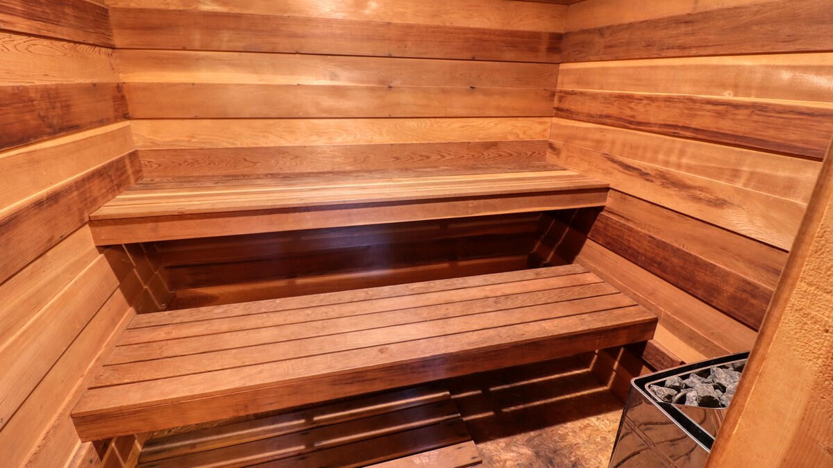 The sauna features wooden benches crafted from warm-toned wood, offering a spacious area for relaxation. The walls are lined with matching wooden planks, creating a cohesive look. A small stove is visible in the corner, designed for heating the space.