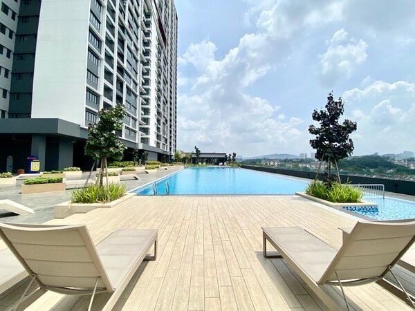 An outdoor swimming pool is showcased, bordered by lush greenery and wooden decking. Comfortable loungers are positioned beside the pool, offering a serene space for relaxation. The backdrop features a modern condominium and a clear sky, enhancing the inviting atmosphere.