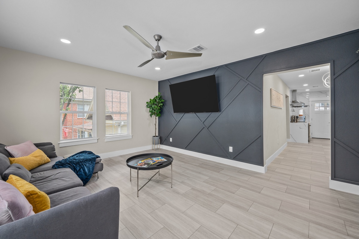 A spacious living room features large windows allowing natural light to fill the area. A modern sectional sofa, adorned with colorful throw pillows, faces a sleek coffee table. A wall-mounted television is visible, with a view leading to the kitchen area beyond.