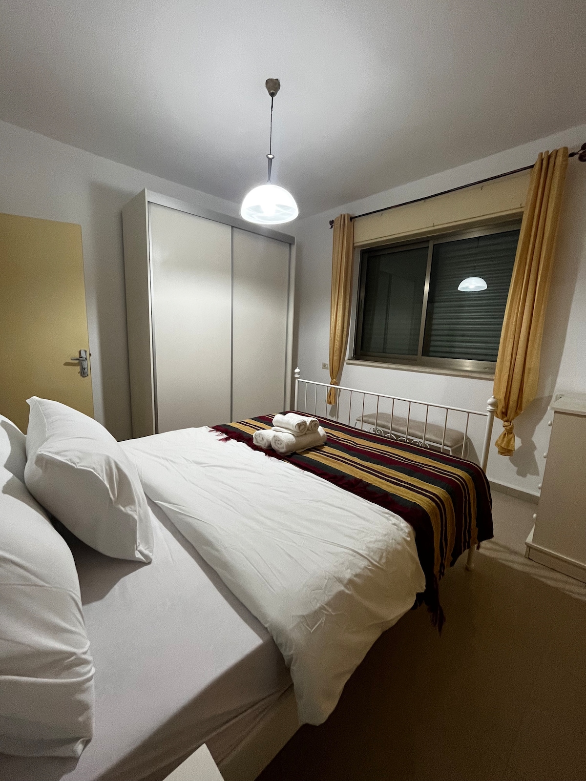 A cozy bedroom features a spacious bed adorned with crisp white linens and decorative pillows. A soft blanket is draped across the foot of the bed. A large closet stands against one wall, while soft yellow curtains frame a window that allows natural light to enter.