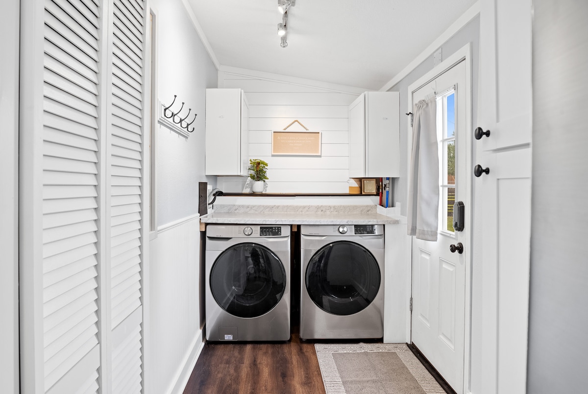 A functional laundry area is presented, featuring two sleek, front-loading washing machines positioned against the wall. Above the machines, cabinetry provides storage, while a small potted plant adds a touch of greenery. The adjacent door leads outside, and hooks for hanging items are mounted on the wall.