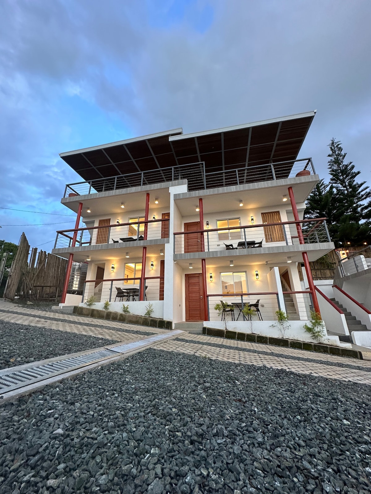 A modern three-story house is viewed from an angle, featuring a spacious balcony with railing. Large windows and sliding doors illuminate the exterior. The surrounding area showcases a landscaped pathway with gravel, adding to the welcoming atmosphere of the property.