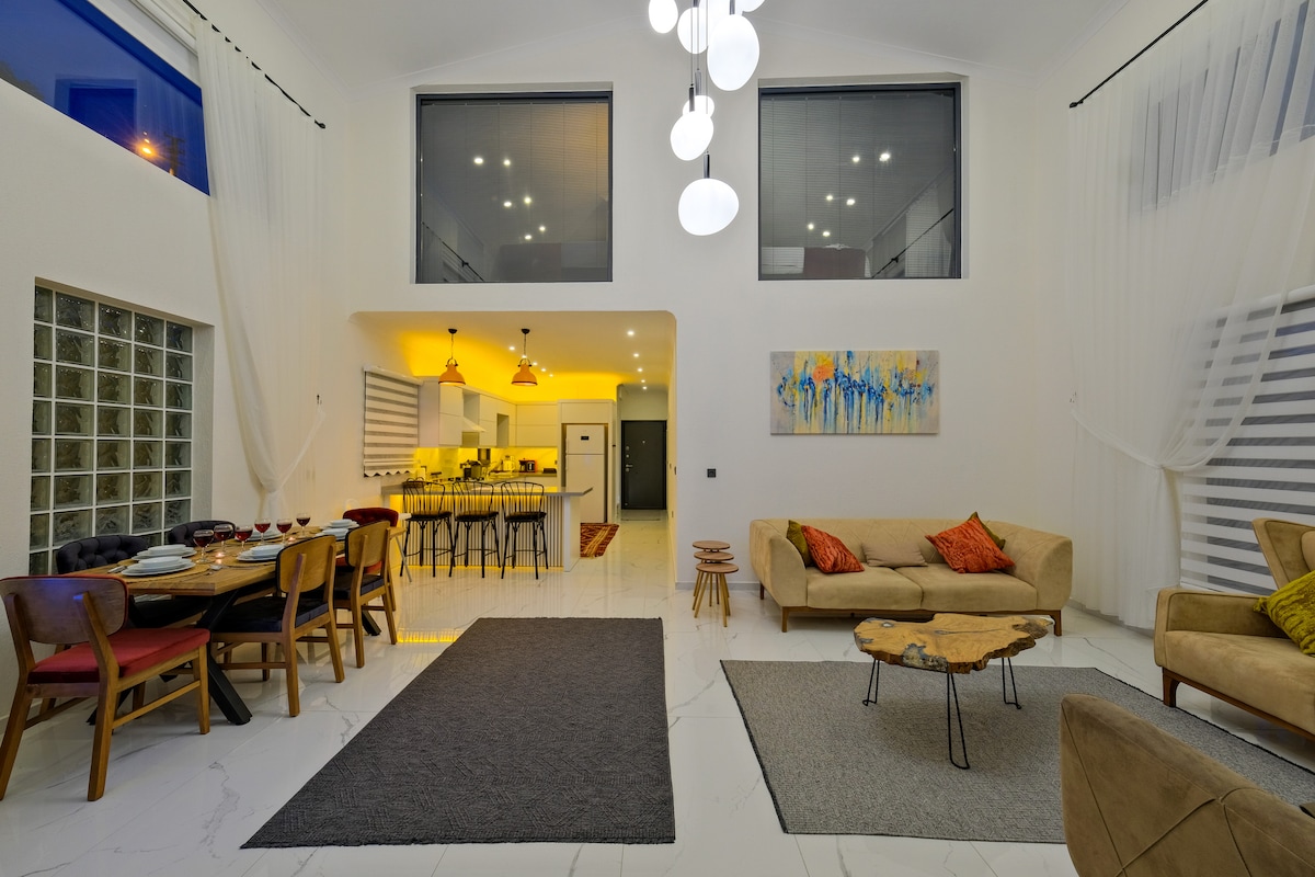 A spacious living area features high ceilings and large windows, allowing natural light to fill the room. A dining table with chairs is positioned alongside a cozy seating area, which includes a low coffee table. Elegant wall art adds color to the neutral palette.