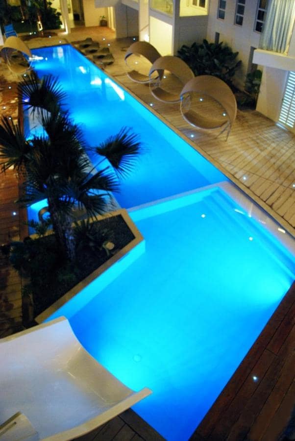 A modern swimming pool is illuminated in soothing blue tones, surrounded by a deck featuring comfortable lounge chairs. Lush greenery enhances the tranquil ambiance, while soft lighting accents the water's surface, providing a serene atmosphere for relaxation.