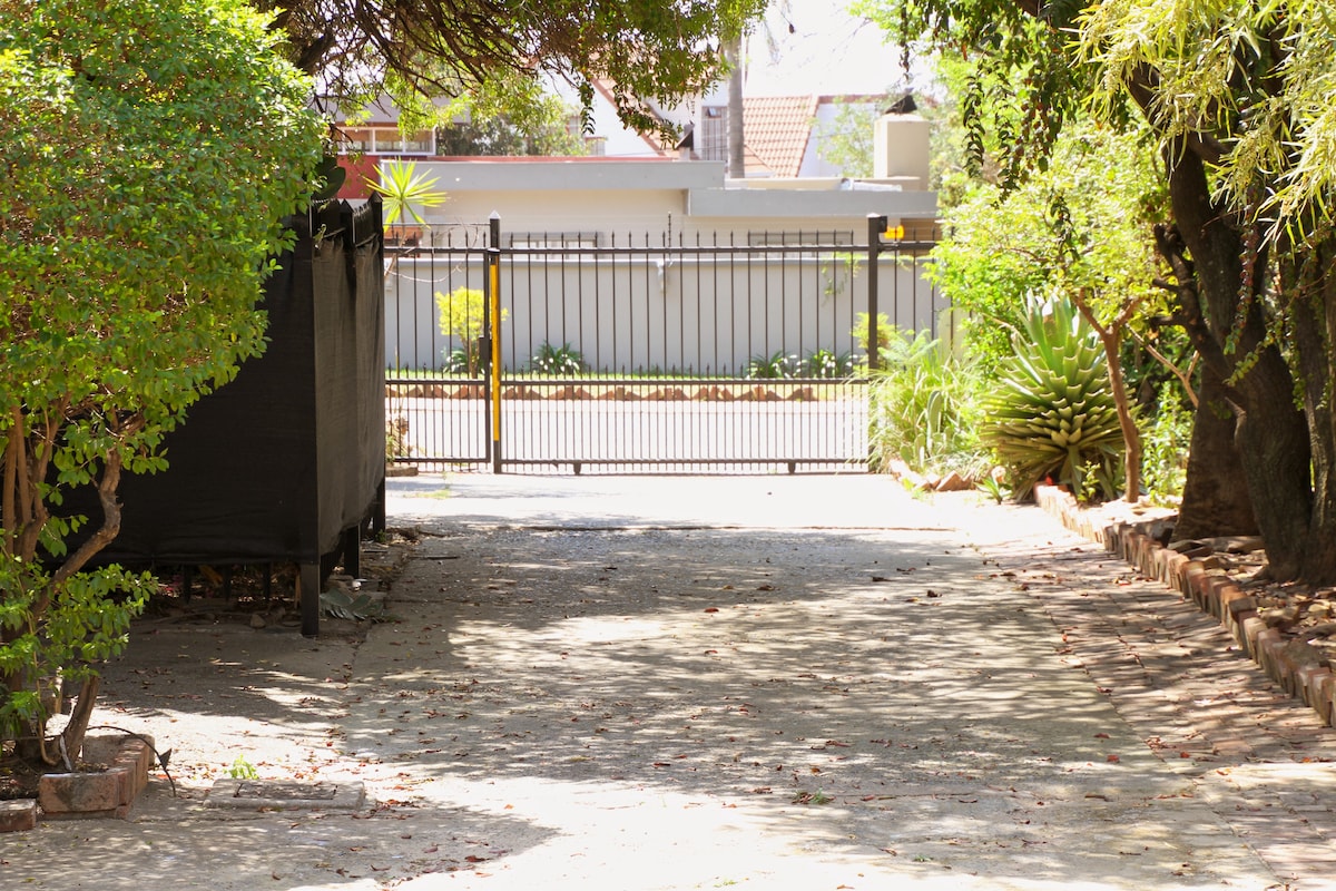 A well-maintained driveway is framed by lush greenery and bushes on either side. A gated entrance is visible in the distance, providing access to the property. Sunlight filters through the trees, illuminating the pathway, which features a combination of paved stones and natural elements.
