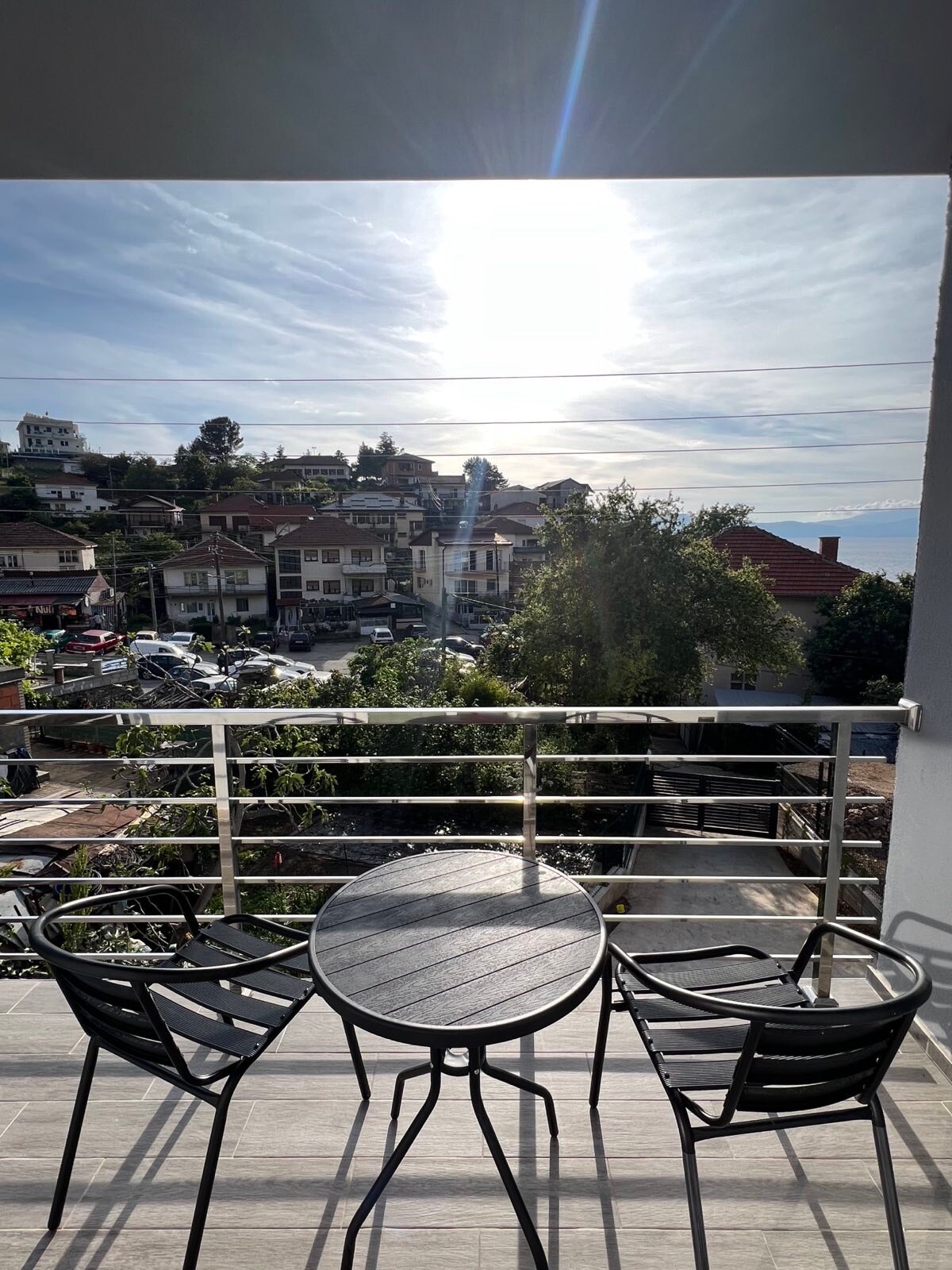 A small balcony is equipped with a round table and two black chairs, offering a vantage point over the village. The sun shines brightly in the sky, illuminating the surrounding buildings and greenery, with a view that extends towards Ohrid Lake in the distance.