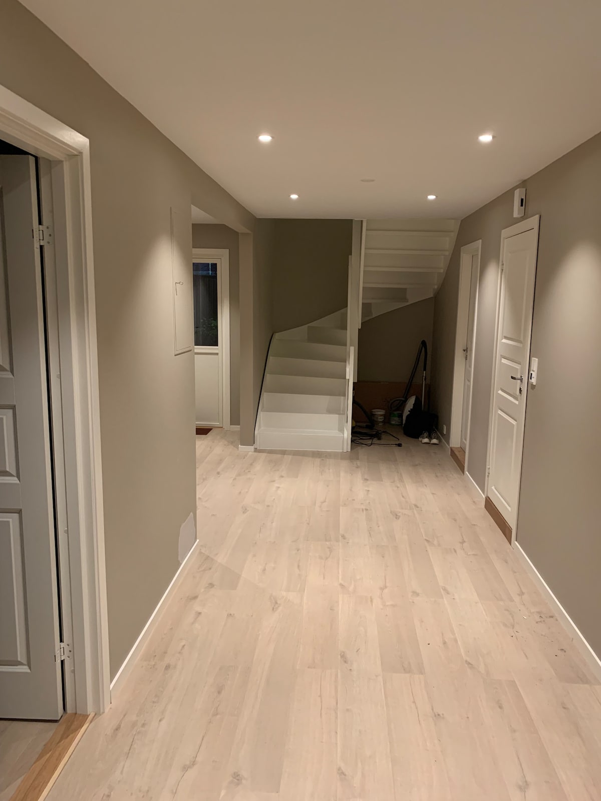 A well-lit hallway features light-colored wooden flooring and neutral-toned walls. Staircase with a white railing leads to an upper level. Several doors are positioned along the walls, providing access to various rooms. Subtle recessed lighting enhances the airy atmosphere.