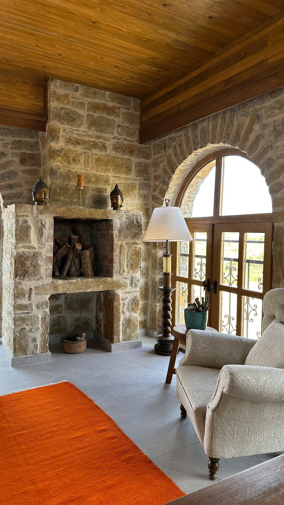 A cozy sitting area is revealed, featuring a soft armchair beside a rustic stone fireplace. Warm wood beams adorn the ceiling, while large arched windows allow natural light to fill the space. An area rug with an orange hue adds a splash of color to the floor.