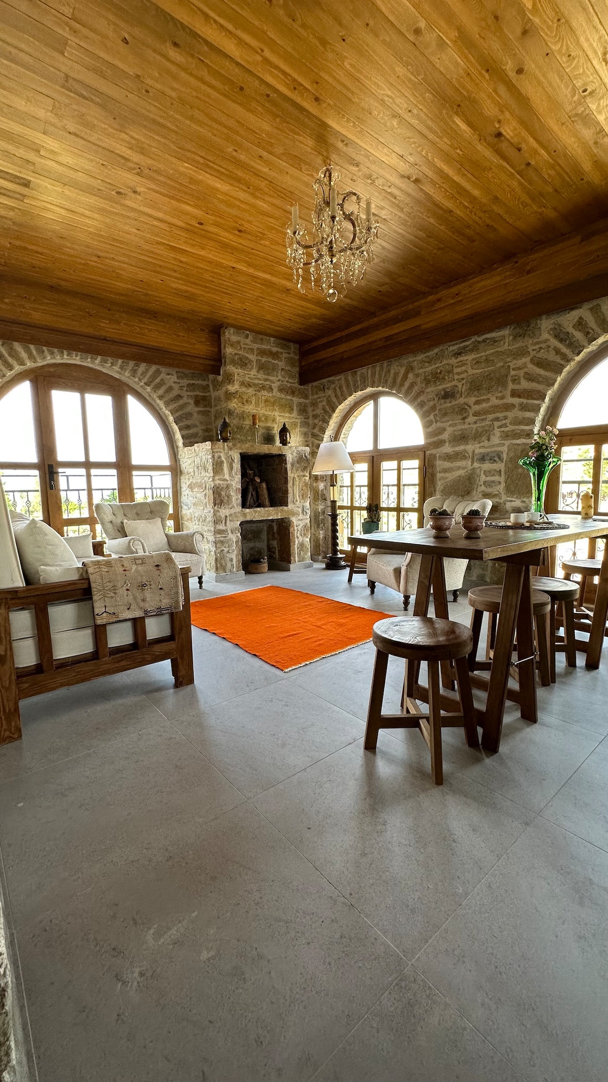 A cozy living space features a wooden ceiling that complements stone walls. A chandelier hangs above a small dining table surrounded by wooden chairs. A vibrant orange rug adds warmth to the area. Large arched windows frame views of the outdoors.