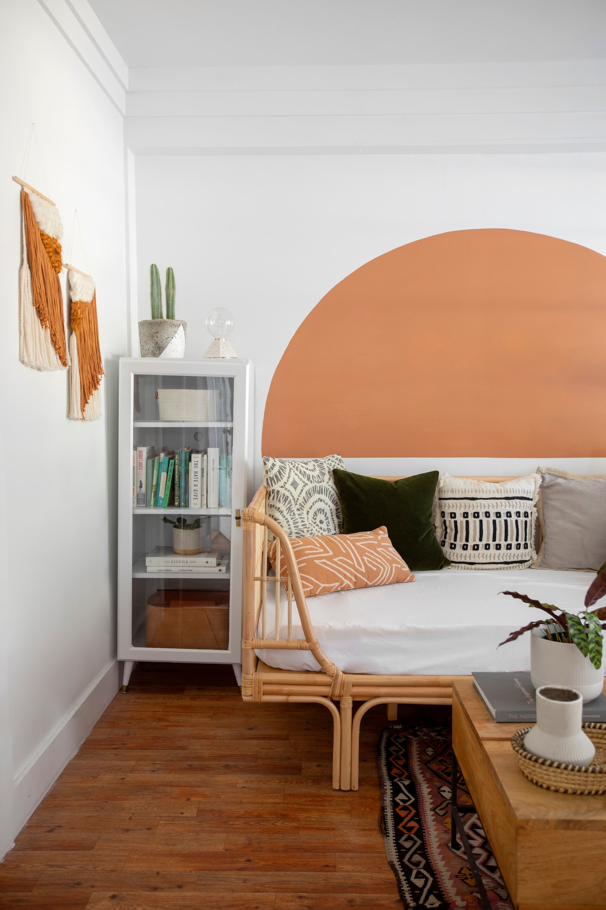 A cozy sitting area features a day bed adorned with an array of textured pillows and a woven throw. A circular terracotta accent is painted on the wall behind, while a glass cabinet displays books and decorative items. Natural light highlights the warm wood flooring.