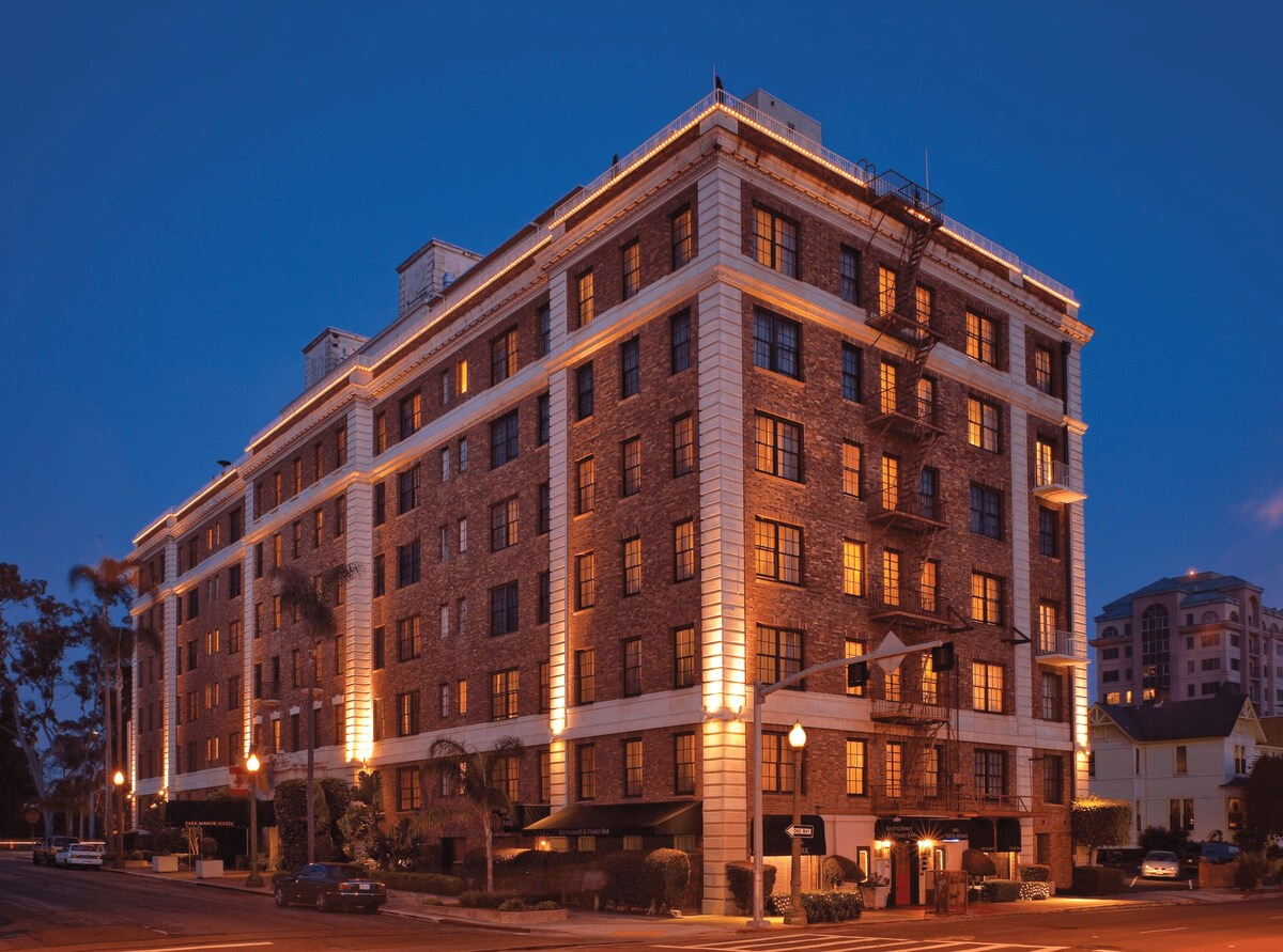 The five-story building features a classic architectural style with stone facades and illuminated windows. Outdoor seating areas are visible at the ground level, complemented by palm trees lining the entrance. Evening lighting enhances the inviting presence of the structure against the twilight sky.