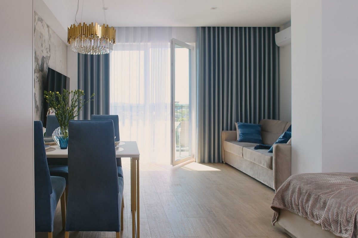 An open-plan living area features a dining table with blue upholstered chairs, complemented by a casual seating area with a light-colored sofa. A large balcony door invites natural light, with sheer curtains adding privacy. Decorative plants accentuate the tabletop, enhancing the space.