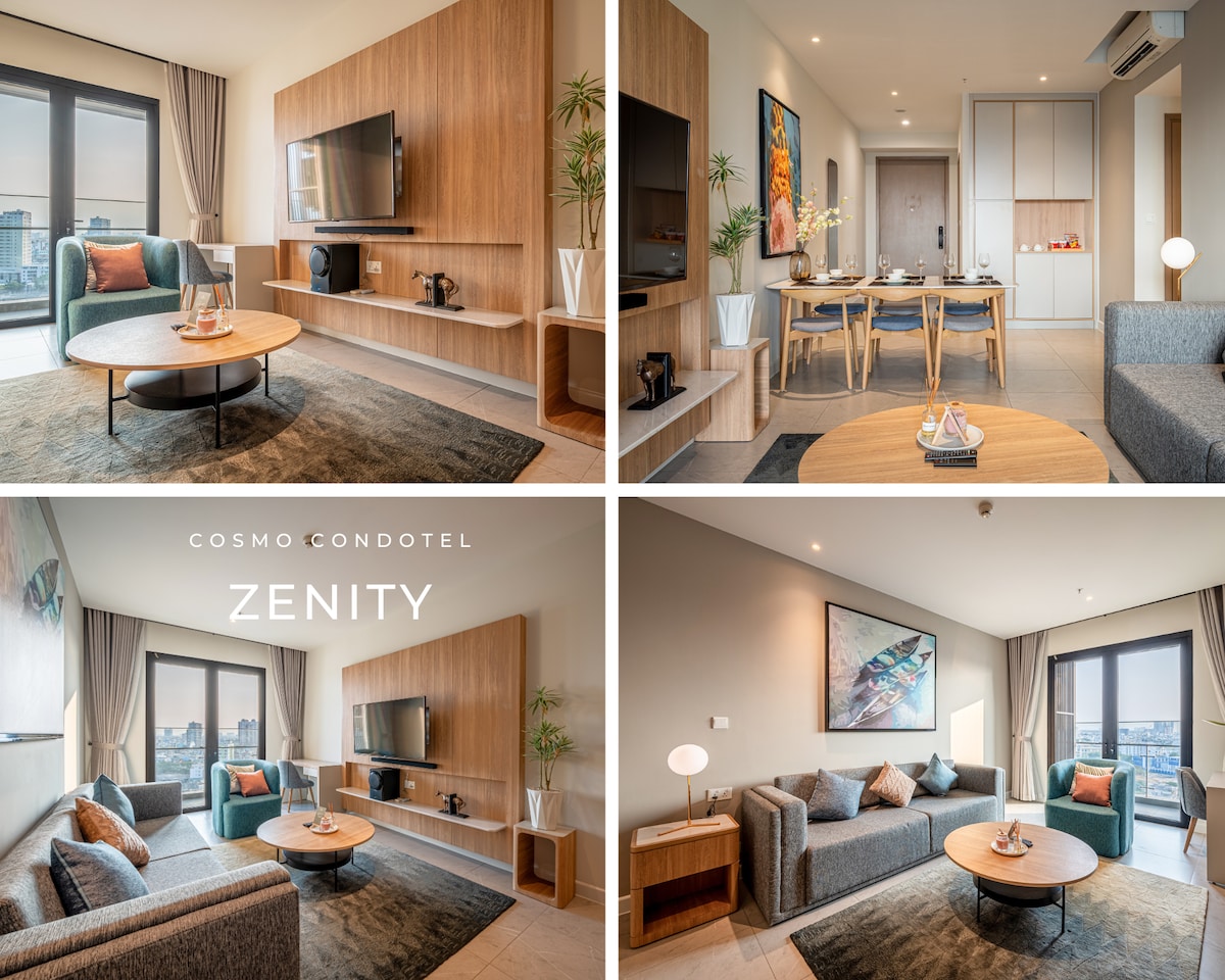 The living area features a soft gray sofa set positioned around a circular wooden coffee table. A wall-mounted TV is present, alongside stylish wooden shelving. A dining table with six chairs is also visible, bathed in natural light from large windows offering a view.