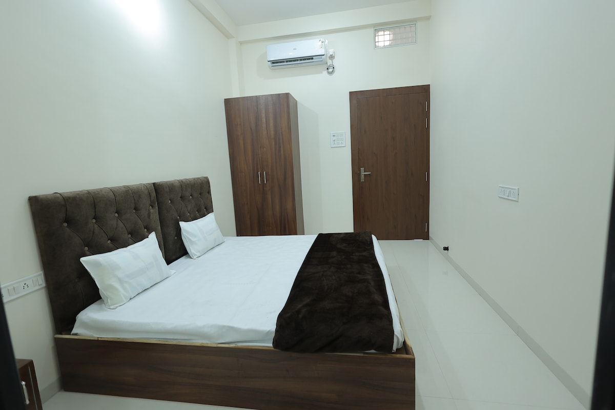 The bedroom features a spacious bed with a soft, dark cover and multiple pillows. Large wardrobes provide ample storage. Air conditioning ensures a comfortable environment, while the minimalist design highlights light-colored walls and polished flooring.