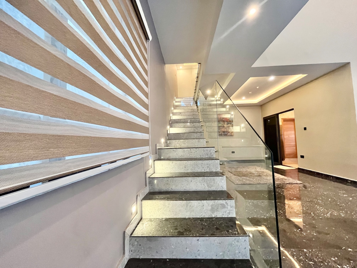 A modern staircase with sleek marble steps leads to an upper level. Natural light filters through a layered window treatment, and subtle lighting illuminates the pathway. The polished floor reflects the elegant design surrounding the stairs, creating a spacious feel.