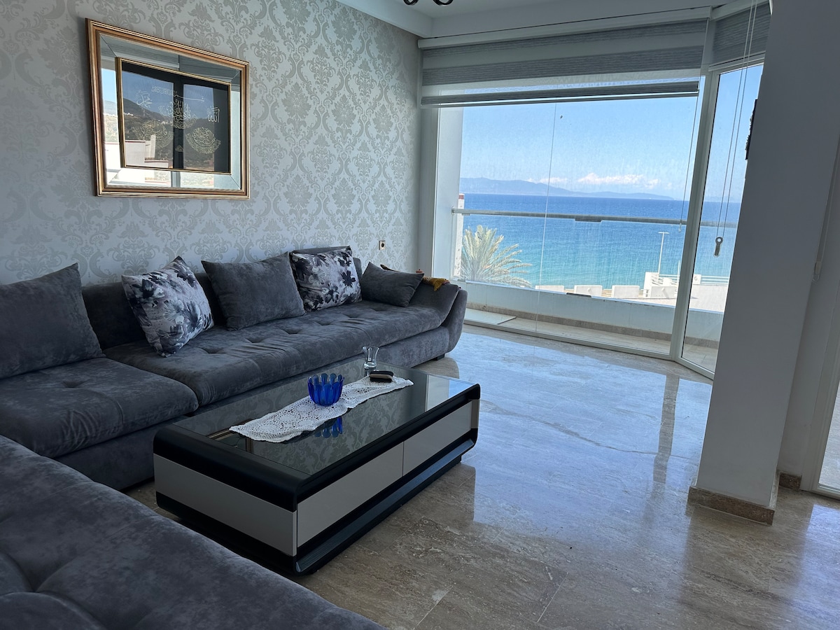 A spacious living area features a large, L-shaped grey sofa facing expansive glass windows. The panoramic view captures the sea and distant mountains. A sleek coffee table with a glass top sits in the center, enhancing the modern aesthetic of the space.