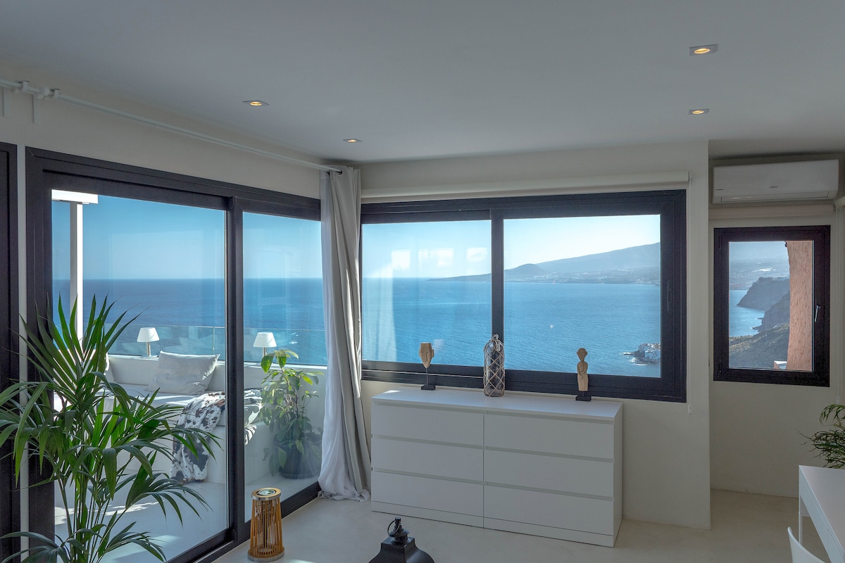 A bright living area features large windows that offer expansive ocean views. A white chest of drawers is situated below the windows, while decorative elements, including plants, enhance the space. Natural light floods the room, creating an open and airy atmosphere.