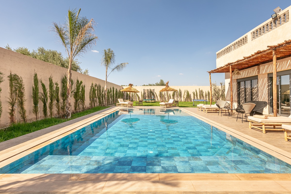 A spacious outdoor area features a large swimming pool with a vibrant blue-tiled surface. Sun loungers surround the pool, and tall palm trees provide a touch of greenery. A shaded seating area is visible, offering a comfortable space to relax while enjoying the serene landscape.