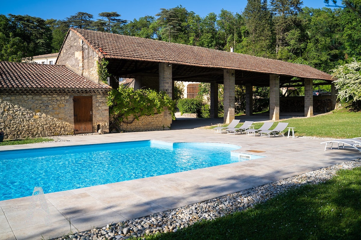 A clear blue pool is set against a rustic stone building with a tiled roof. Lounge chairs are positioned around the pool area, which is surrounded by manicured grass and trees. Natural light enhances the serene outdoor setting.