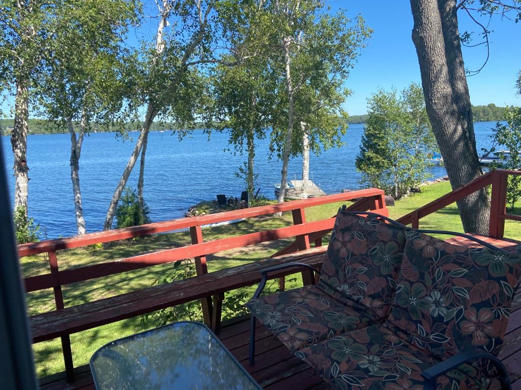 A deck furnished with floral-patterned seating overlooks the serene waters of Lake Nosbonsing. Lush greenery surrounds the area, with trees providing shade. The natural landscape leads to the private waterfront, enhancing the view of the lake and distant shoreline.