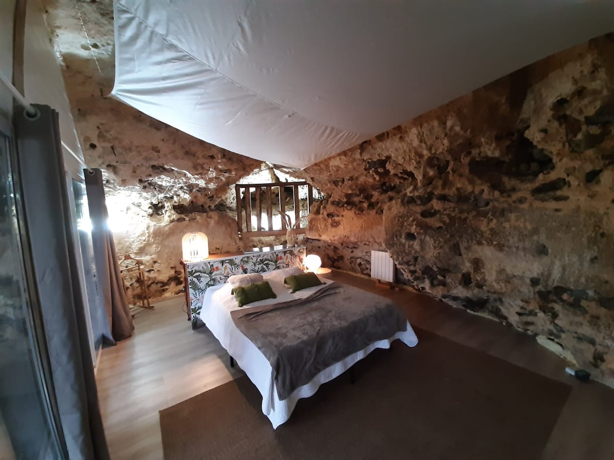 A unique bedroom features textured stone walls and a vaulted ceiling. A spacious bed is surrounded by soft lighting from lanterns, with decorative pillows positioned neatly. Natural light filters through a small window, providing a warm ambiance within the rustic setting.