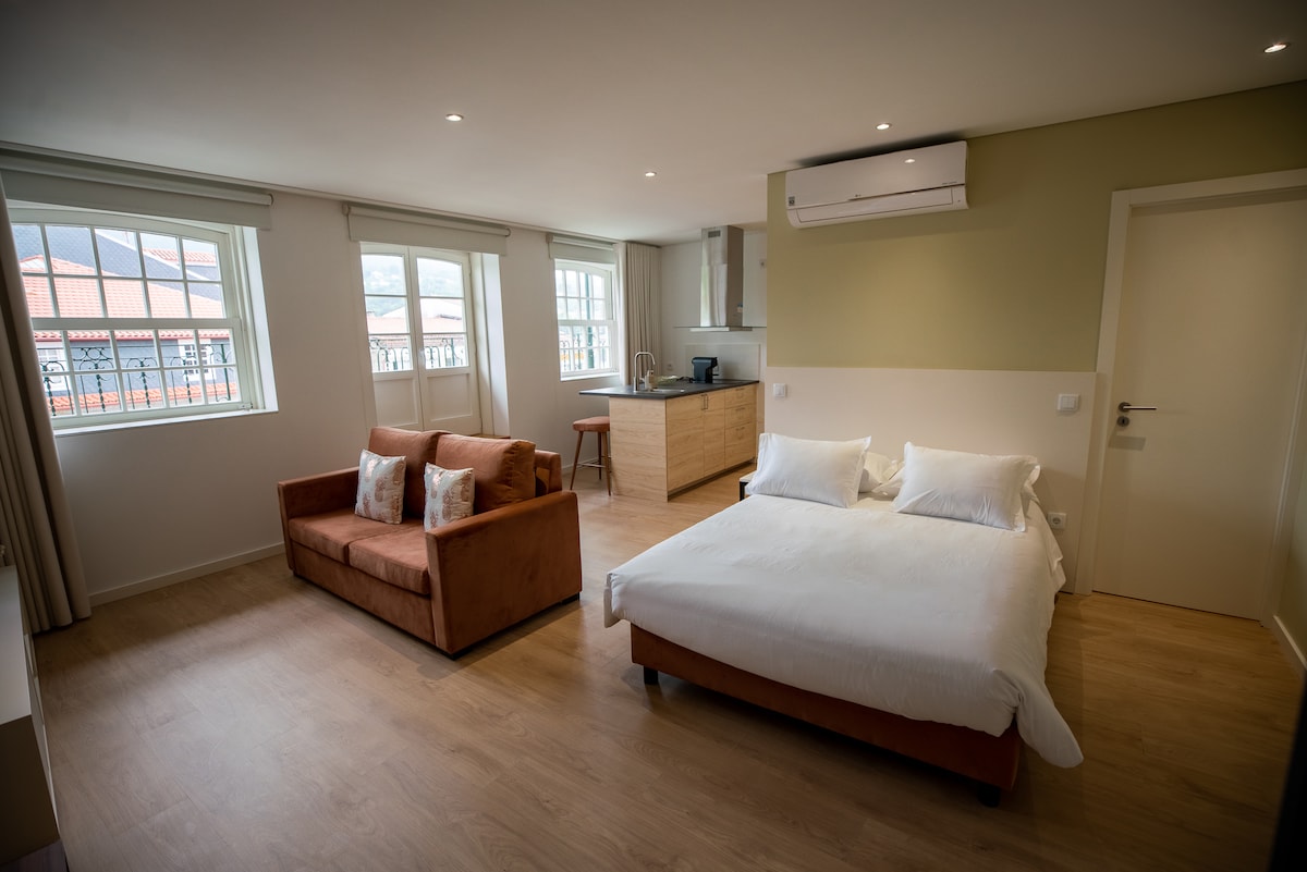 A bright studio apartment is showcased with large windows providing natural light. A spacious bed is positioned beside a comfortable couch. A kitchenette is visible in the background, equipped with essential appliances. Neutral tones and modern furnishings create a welcoming environment.