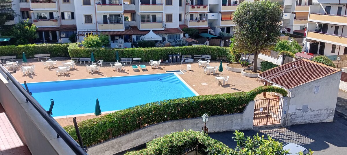 An outdoor swimming pool is surrounded by neatly arranged sun loungers and umbrellas. Lush greenery encloses the pool area, offering privacy. The space is well-maintained, with a pathway leading to the entrance, creating an inviting atmosphere for relaxation.