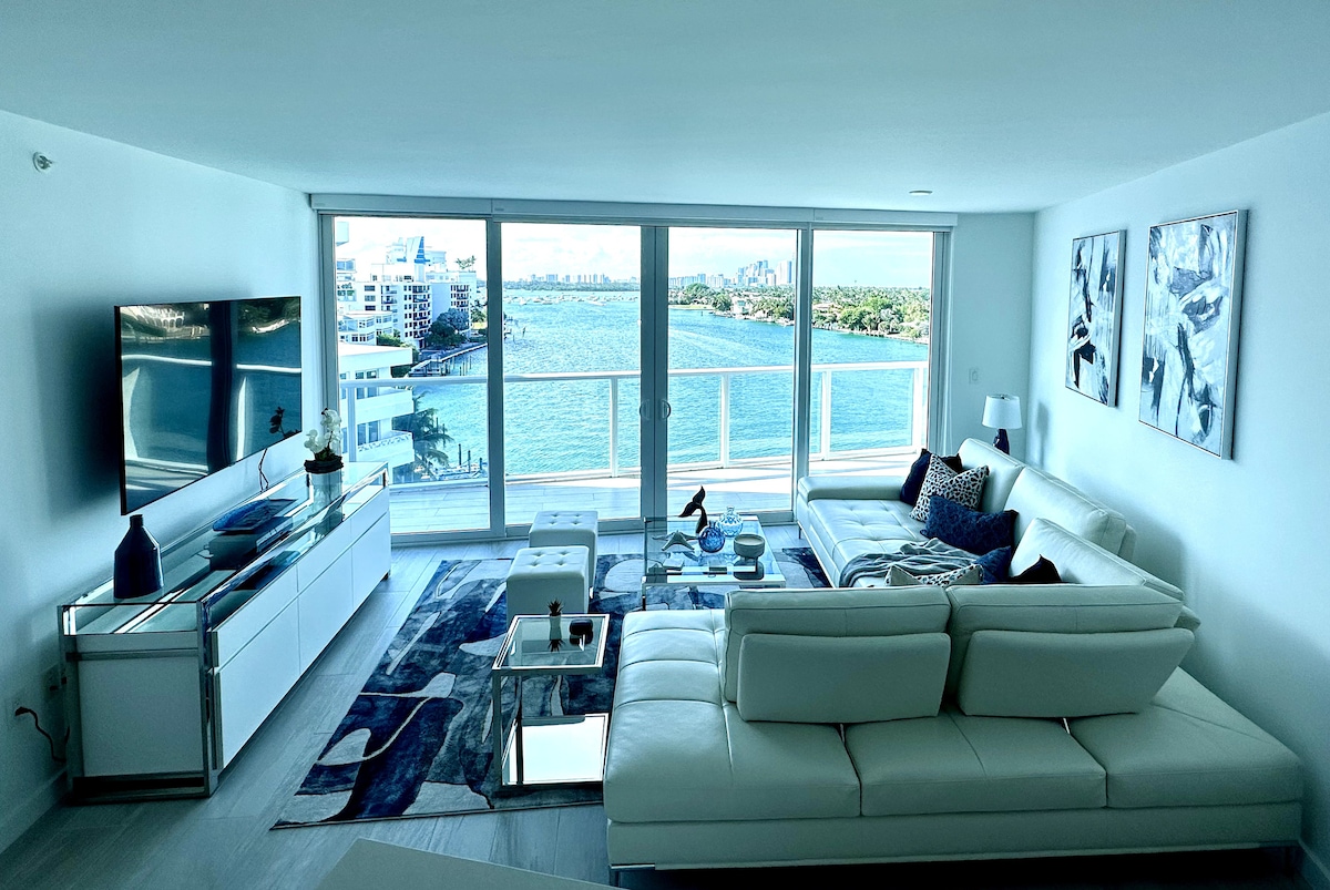 A modern living area is showcased with large glass doors framing views of the water and skyline. A white sectional sofa complements a sleek coffee table, while a television is mounted on a white console. Artwork adorns the walls, enhancing the spacious feel.