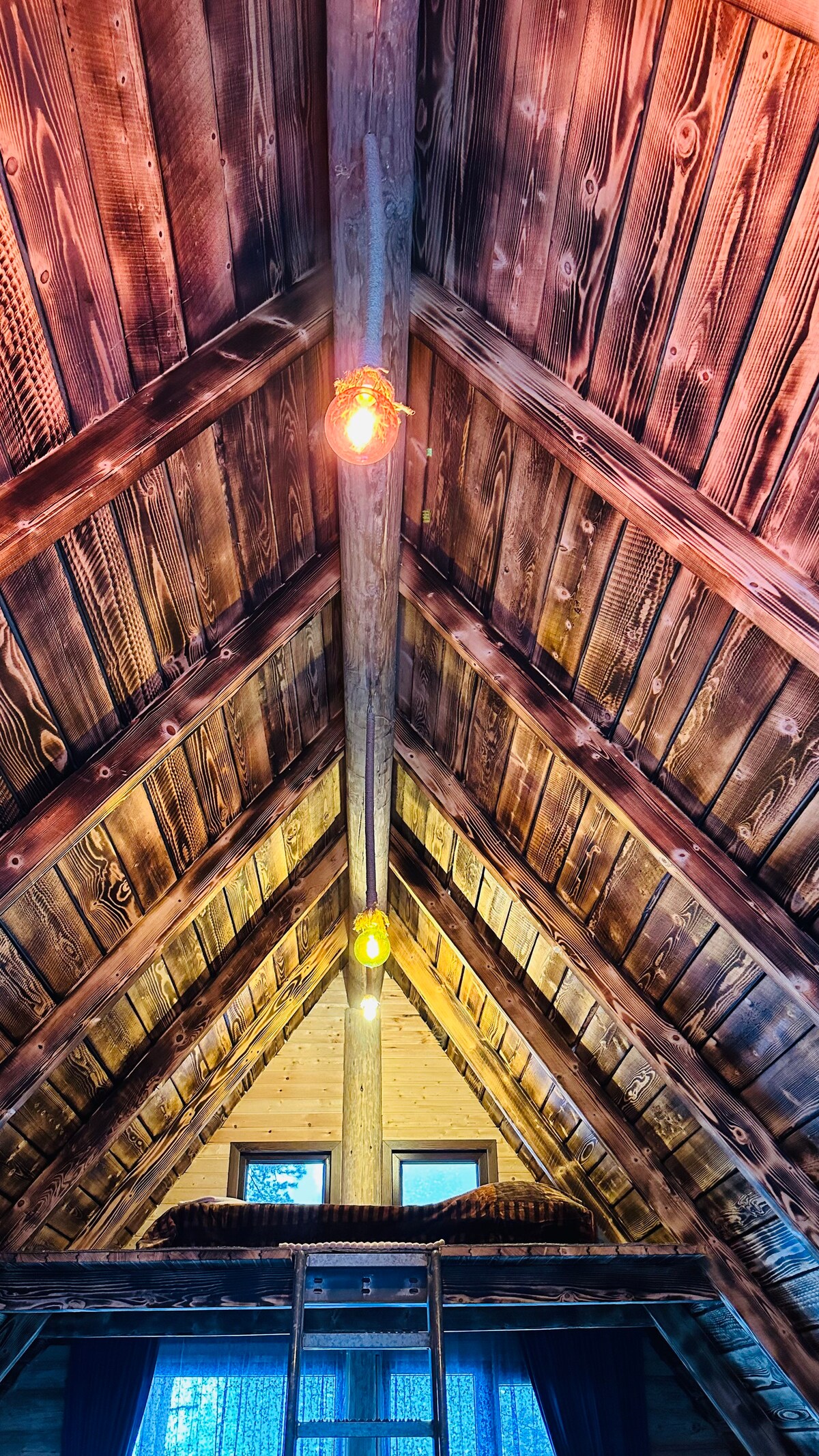 The interior of the cottage features a vaulted wooden ceiling, showcasing rich timber textures. Soft lighting is provided by pendant fixtures hanging from the beams. A wooden ladder leads to a sleeping area, with windows offering views of the surrounding forest.
