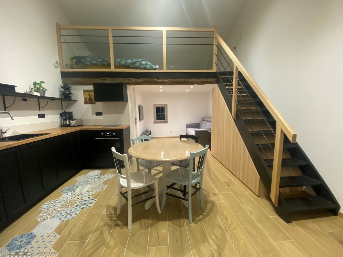 An open living space is shown, featuring a wooden dining table with four chairs and a compact kitchen area. A staircase leads to a mezzanine with a spacious bed. Soft lighting enhances the modern design and light wood flooring throughout.