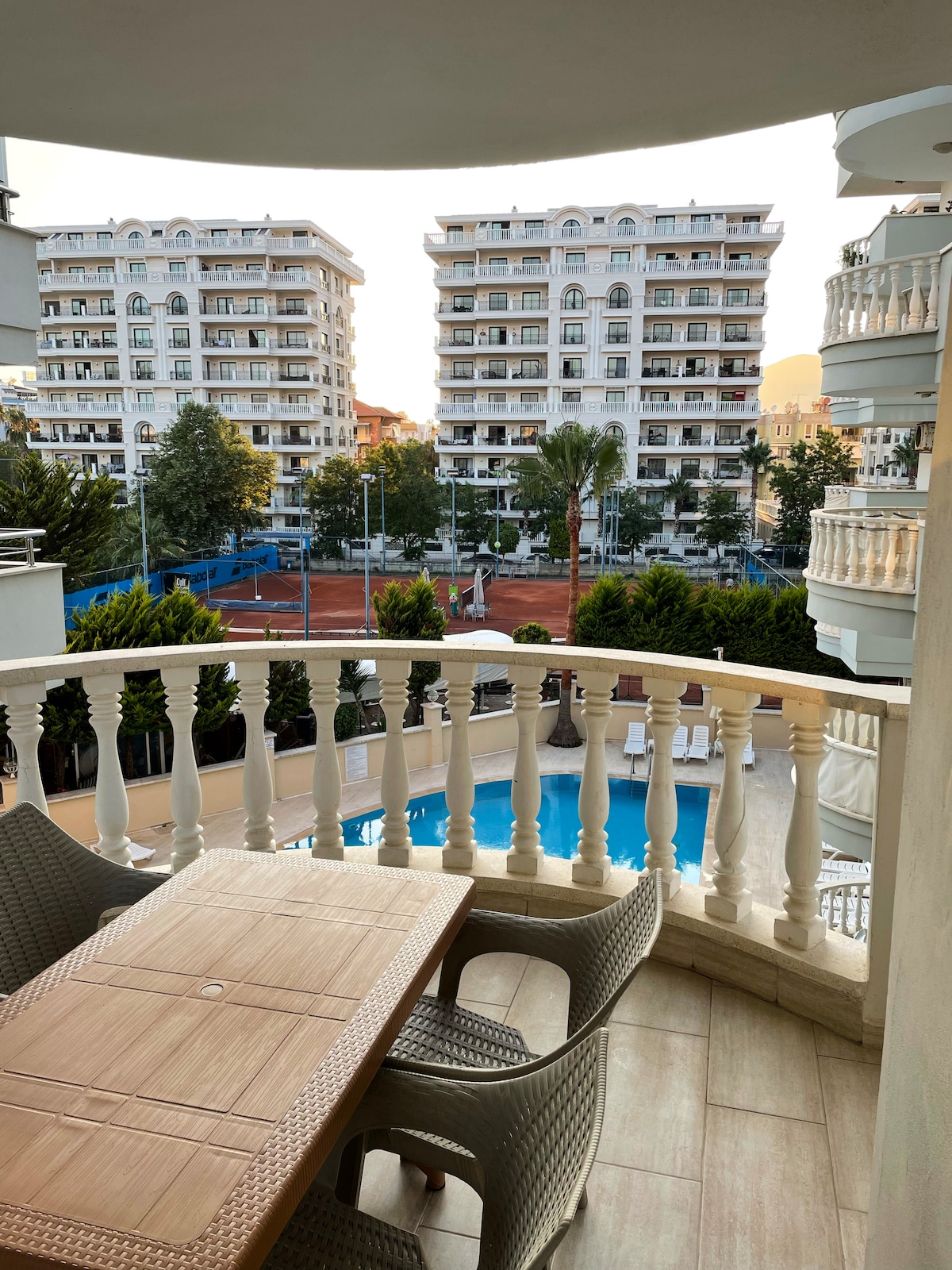 A balcony view features a wooden table with two chairs, overlooking a swimming pool surrounded by greenery. Multi-story buildings are visible in the background, alongside a recreational area with tennis courts. Natural light brightens the scene as evening approaches.