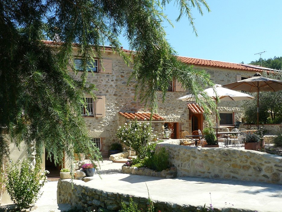 A rustic stone building is surrounded by lush greenery, featuring a red-tiled roof. Outdoor seating with umbrellas is visible on a raised terrace, along with well-maintained flower beds, creating a tranquil outdoor space. Sunlight casts a warm glow across the scene.