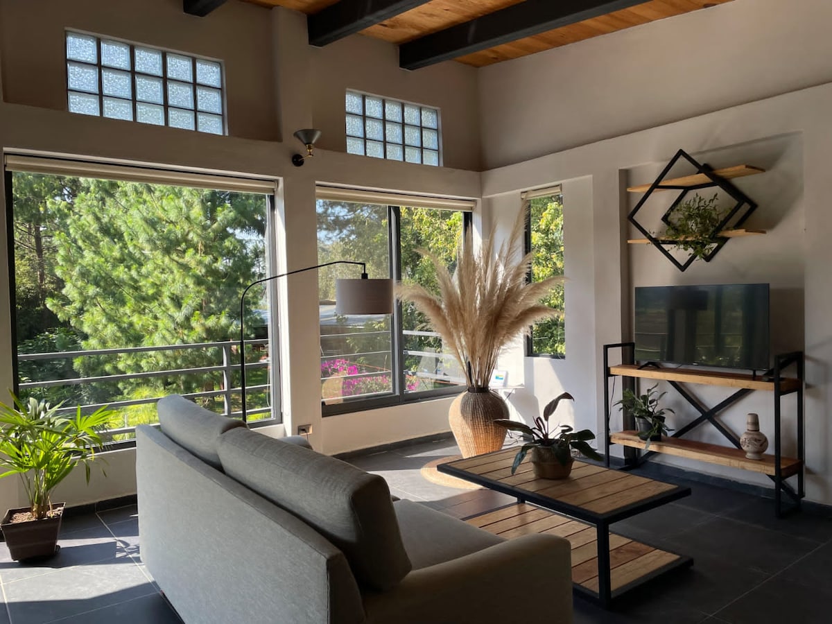 A well-lit living area features large windows that provide views of lush greenery. A comfortable sofa faces a modern TV stand, while a decorative vase with tall pampas grass adds a touch of nature. Potted plants enhance the serene ambiance.