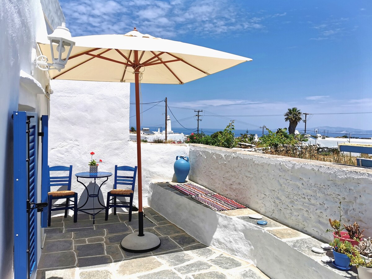 An outdoor space features a round table and two blue chairs, shaded by a large umbrella. A decorative rug lies on a stone bench, with a blue ceramic pot and a flower pot adding color. A glimpse of the sea and palm trees can be seen in the background.