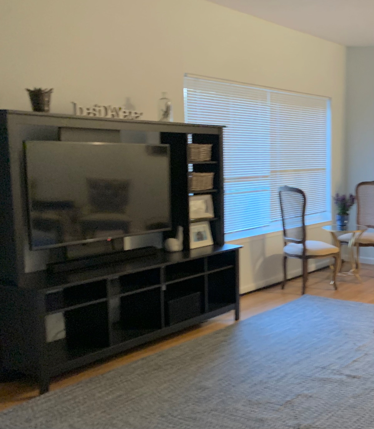 A welcoming living area features a dark entertainment unit with a flat-screen television and decorative items. Two light-colored chairs sit near a window, allowing natural light to fill the space. A textured area rug covers the wooden floor, adding warmth to the room.