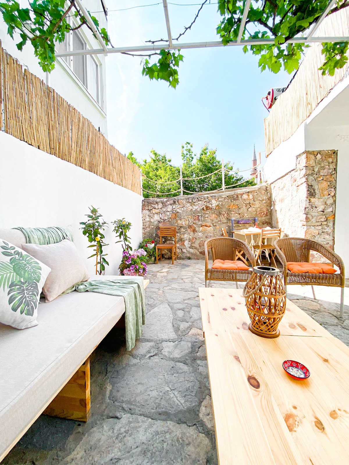 A charming outdoor garden area features a stone-paved floor, surrounded by greenery and flowering plants. Comfortable seating is placed alongside a wooden table, creating a welcoming space for relaxation. Natural light filters through the overhead vines, enhancing the serene atmosphere.
