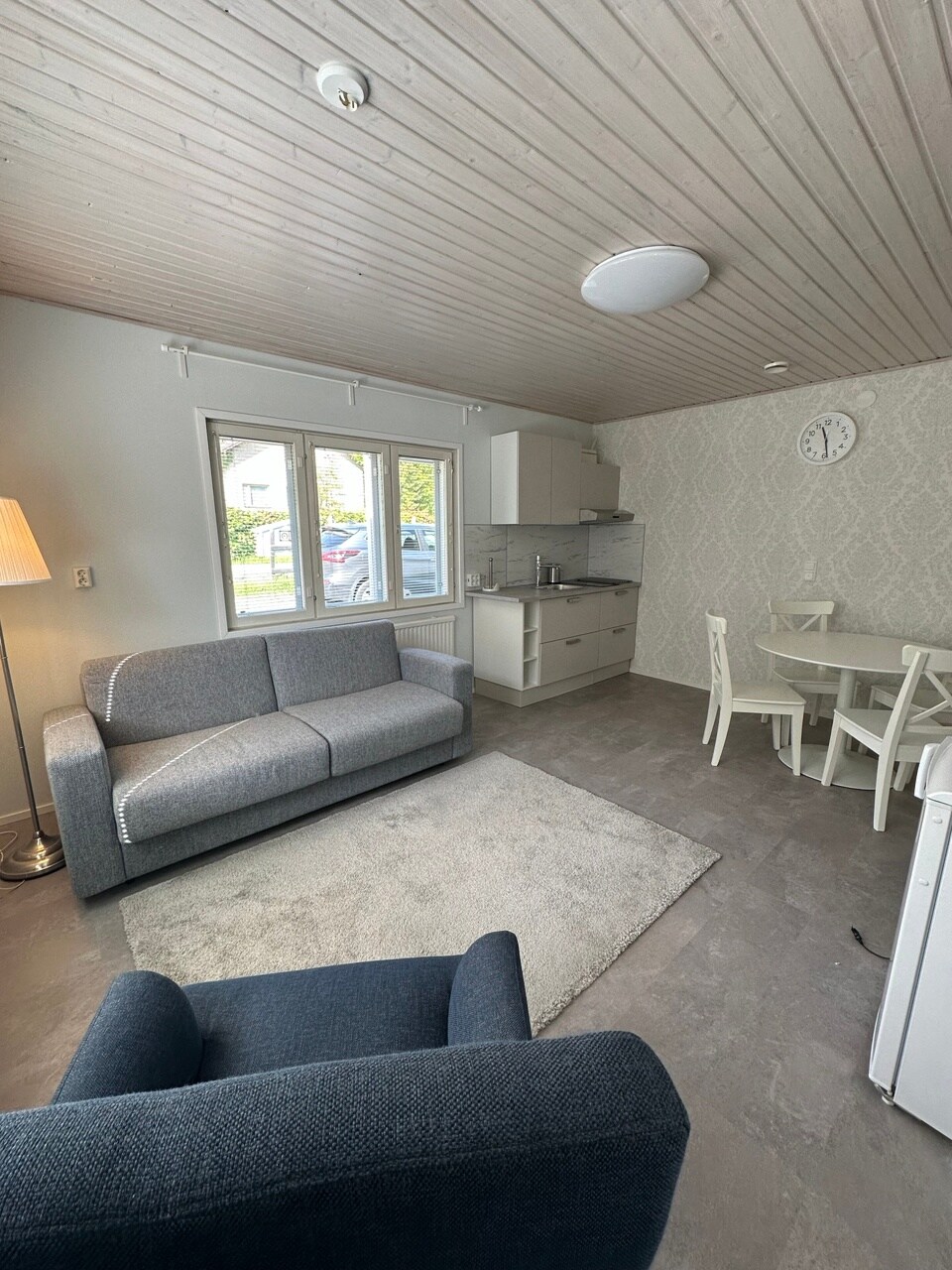 A cozy living area features a grey sofa and a blue armchair situated on a soft rug. A round dining table with white chairs is visible, along with a well-equipped kitchen. Natural light enters through two large windows, highlighting the light-colored walls and ceiling.
