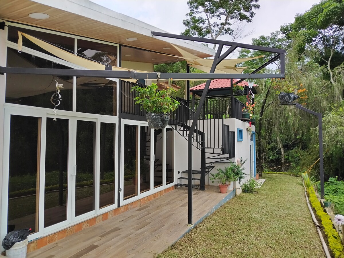 The exterior features a modern design with large glass panels, providing an abundance of natural light. An exterior staircase leads to a second level, while hanging planters add greenery. A spacious deck area is positioned adjacent to the well-maintained lawn.