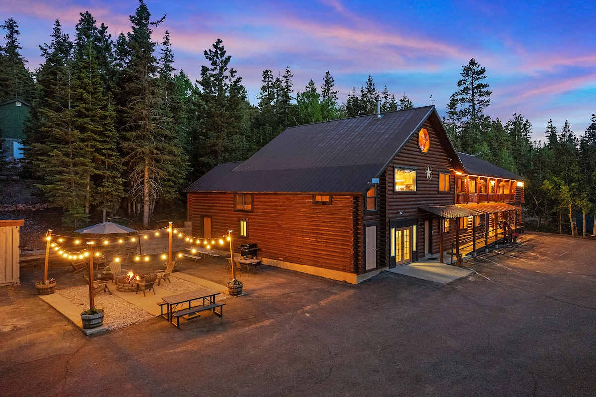 The exterior of a spacious log cabin features a large outdoor area with string lights illuminating a barbecue and firepit setup. The building is surrounded by tall trees, with large windows reflecting the twilight sky.